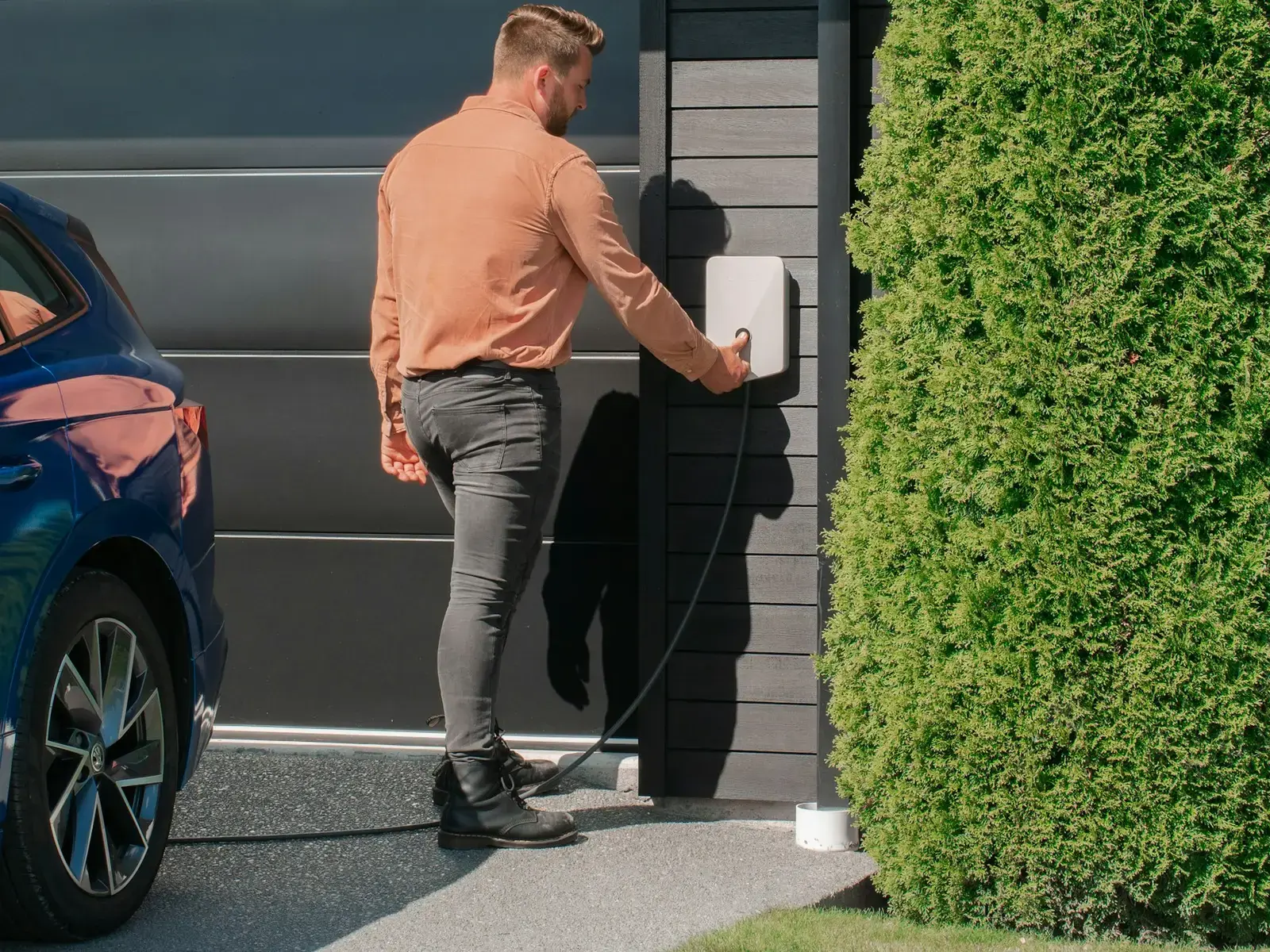 Un homme branche le câble de recharge sur une borne murale, devant sa maison, à côté de sa voiture électrique bleue.