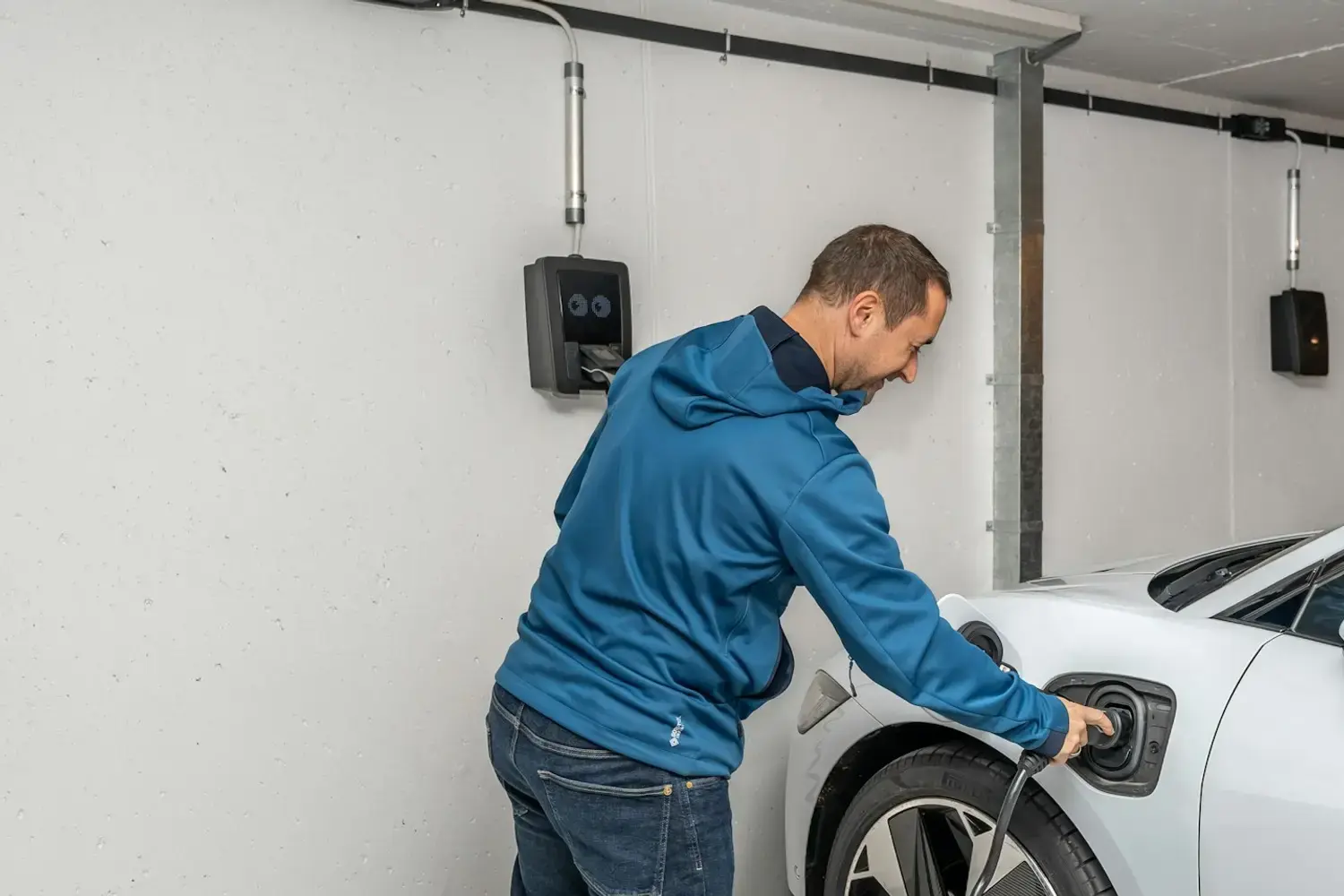 Un homme branche le câble de recharge sur sa voiture électrique blanche, dans un garage équipé d'une borne murale.