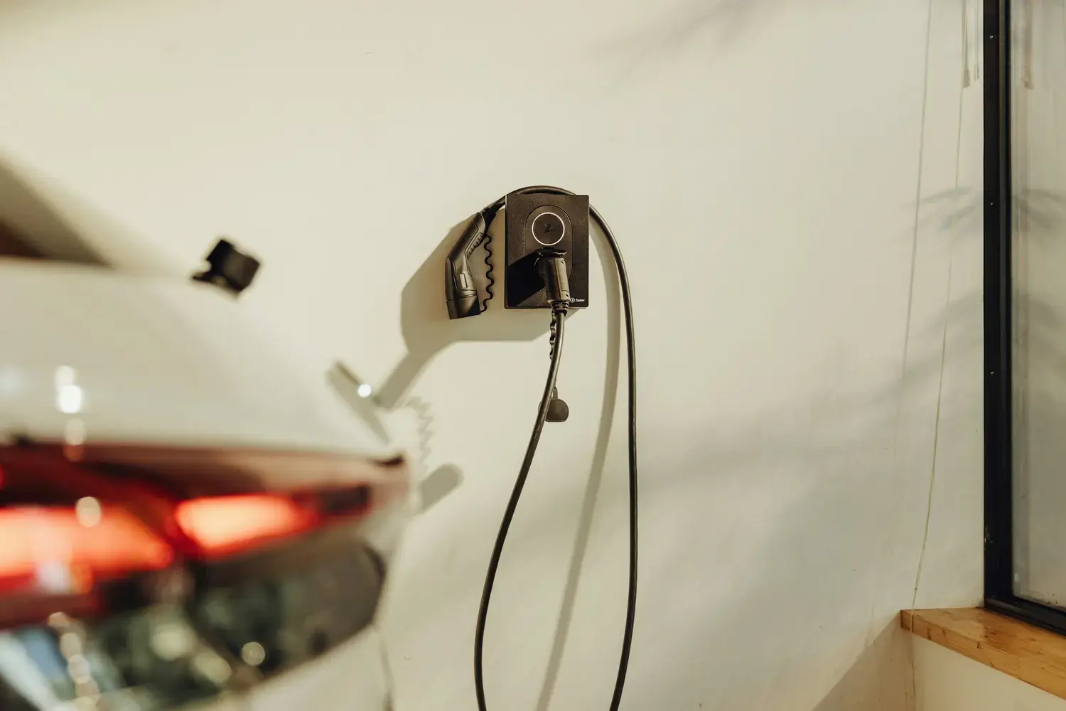 Une borne de recharge murale fixée au mur clair d'un garage, son câble enroulé autour du boîtier, l'arrière d'une voiture électrique à côté.