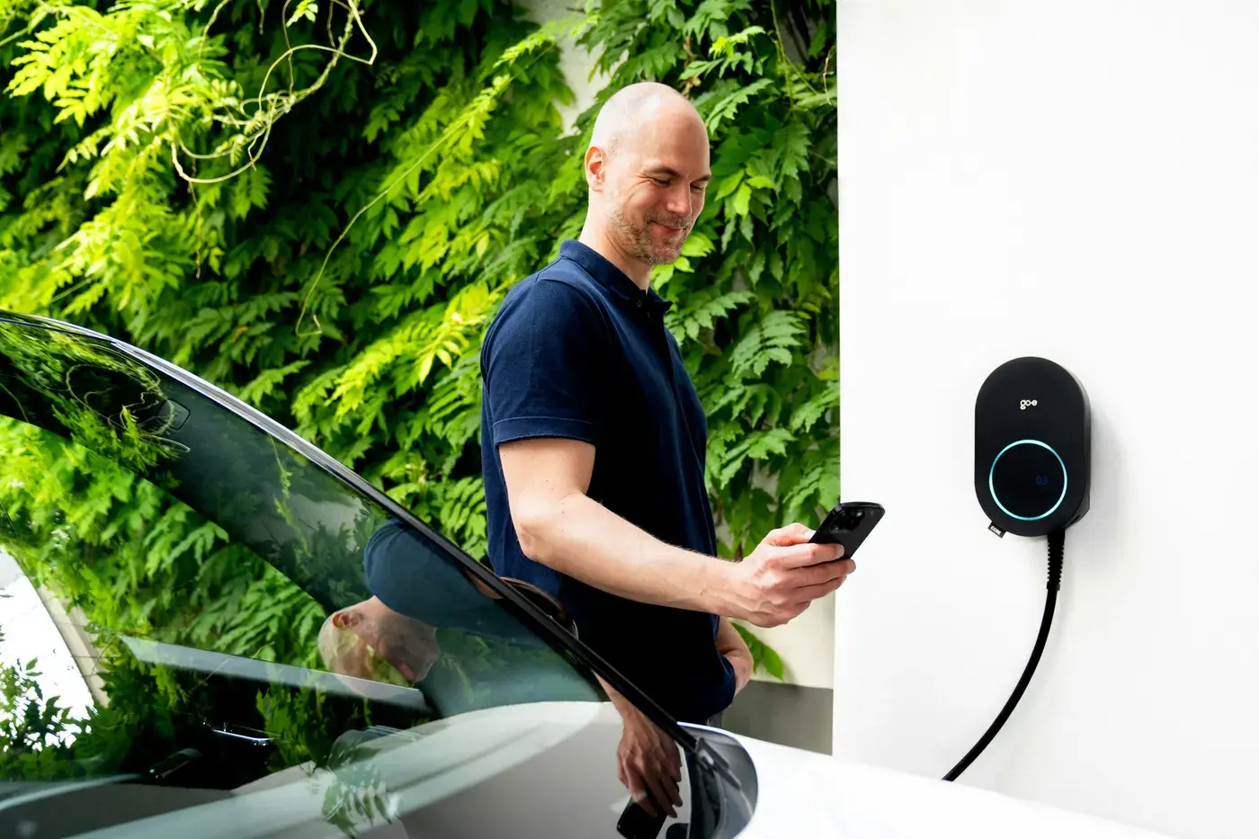 Un homme consulte son téléphone près de sa voiture électrique, à côté d'une borne de recharge murale fixée sur un mur blanc.