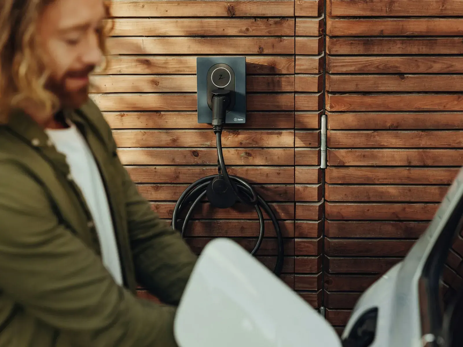 Une borne de recharge murale et son câble lové sur un bardage en bois, devant une voiture électrique blanche.