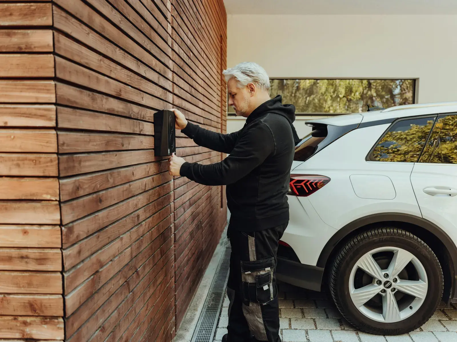 Un homme en vêtements de travail intervient à deux mains sur une borne de recharge murale, à côté d'une voiture électrique.
