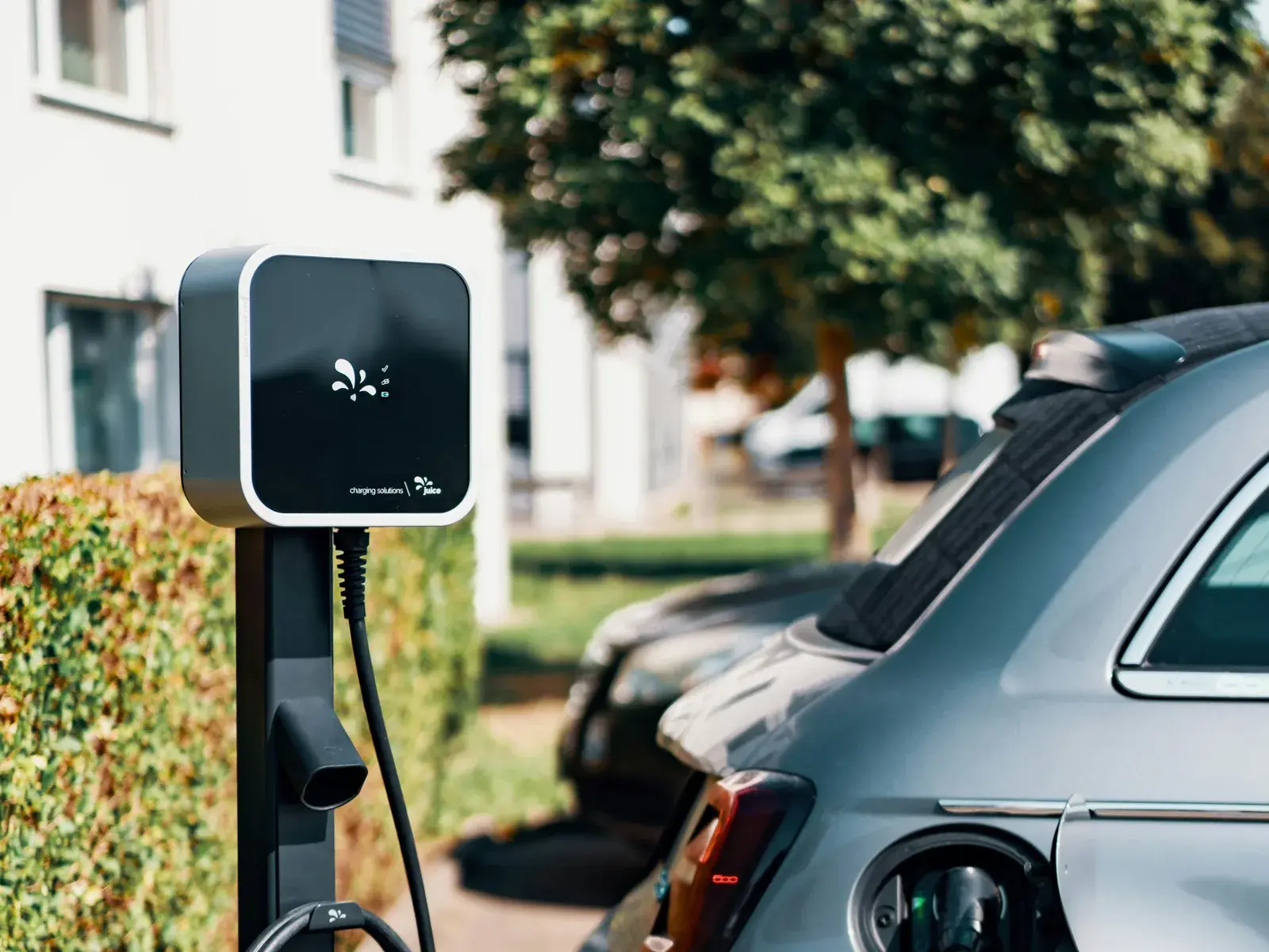 Une borne de recharge sur pied, son câble branché à la trappe de charge d'une voiture stationnée le long d'une haie.